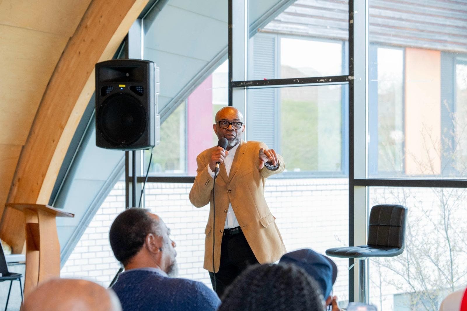 Patrick Burrows in a tan blazer holding a microphone and pointing, speaking in a modern glass-walled room.